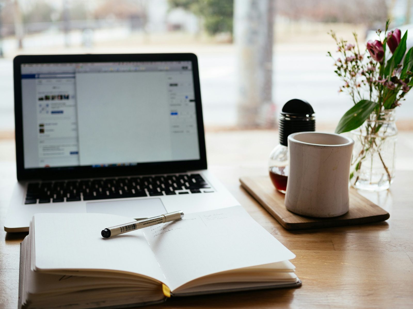 A workspace with a laptop, an open notebook with a pen on it, a coffee mug, a glass jar with flowers, and a tray with a glass teapot and a black container, set on a wooden table near a large window.