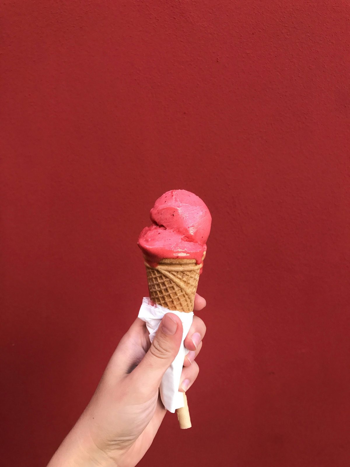 Hand holding a pink ice cream cone with a swirl of scoop on a waffle cone, against a red background.