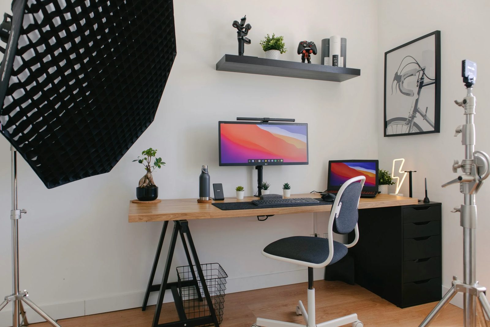 Home office setup with a wooden desk, dual monitors, potted plants, a laptop, desk lamp, and photo frame, with studio lighting equipment in a bright room