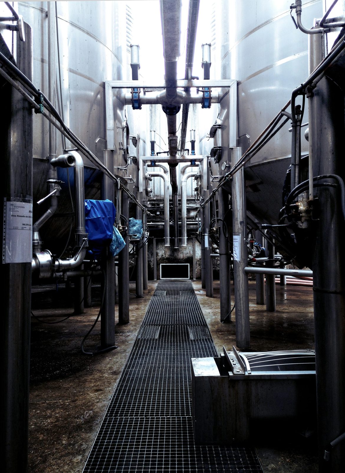 A stainless steel industrial brewing or processing tank setup with pipes, valves, and a grated walkway in an enclosed facility.